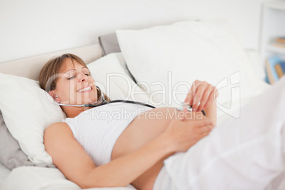 Good looking pregnant woman using a stethoscope while lying on a