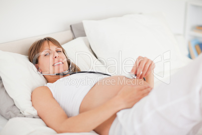 Charming pregnant woman using a stethoscope while lying on a bed