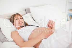 Charming pregnant woman using a stethoscope while lying on a bed