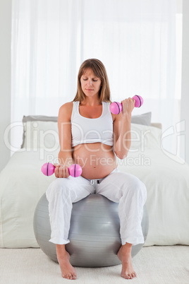 Pretty pregnant female using a dumbbell while sitting on a gym b