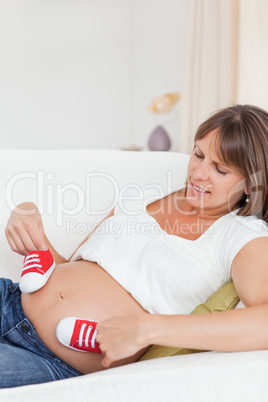 Pretty pregnant female playing with red baby shoes while lying o
