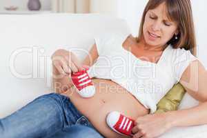 Beautiful pregnant female playing with red baby shoes while lyin