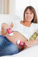 Attractive pregnant female playing with red baby shoes while lyi