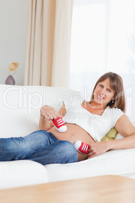 Cute pregnant female playing with red baby shoes while lying on