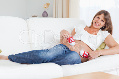Charming pregnant female playing with red baby shoes while lying