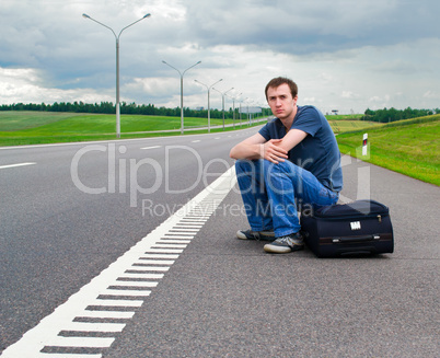 The young man sits pending on road with a suitcase