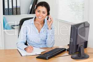 Charming woman using her mobile phone while writing on a sheet o