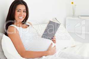 Beautiful pregnant woman reading a book while lying on a bed