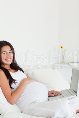 Cute pregnant woman relaxing with her laptop while lying on a be