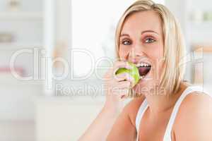 Young blonde woman sitting at table eating a green apple while l