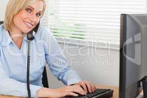 Smiling blonde businesswoman on the phone while typing looks int