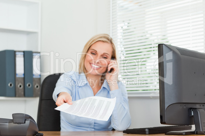 Smiling businesswoman phoning and passing a paper looks into cam