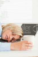 Gorgeous blonde woman sleeping on her notebook holding cup of co