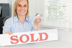Blonde businesswoman showing miniature house and SOLD sign looki