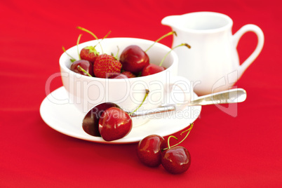 milk jug cup saucer spoon cherry and strawberry on a red backgro