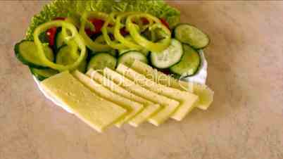 Young man on kitchen table knifes cheese also puts it on plate with tomatoes and cucumbers.