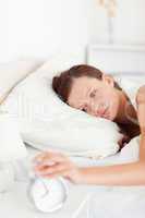 Sleepy red-haired woman lying in bed turning off alarm clock
