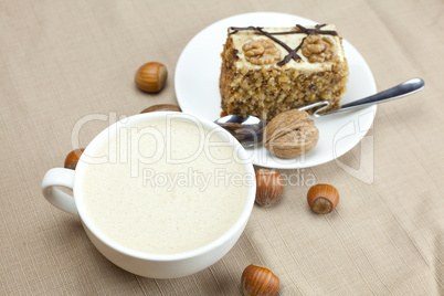 A cup of cappuccino, a piece of cake with nuts and spoon lying o