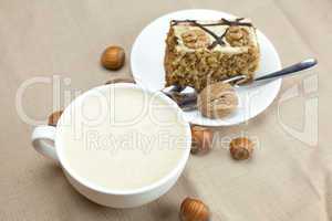 A cup of cappuccino, a piece of cake with nuts and spoon lying o