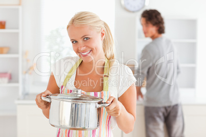 Woman posing with a boiler while her fiance is washing the dishe