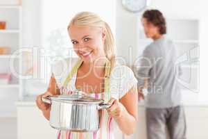 Woman posing with a boiler while her fiance is washing the dishe