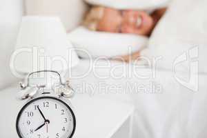 Woman hidding her head in a pillow while the alarrmclock is ring