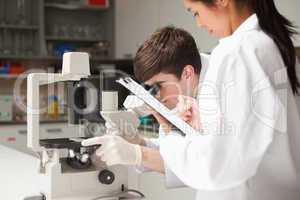 Scientist looking in a microscope while his coworker is writing