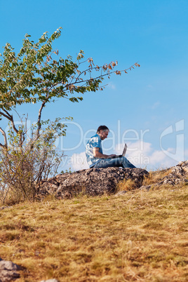 young man using laptop sitting on a hill against the blue sky