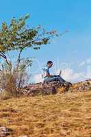 young man using laptop sitting on a hill against the blue sky