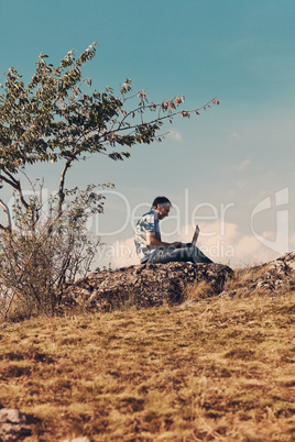 young man using laptop sitting on a hill against the blue sky