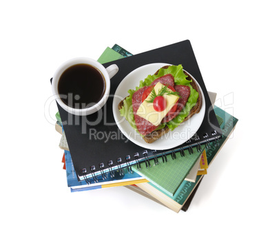 A stack of books and notebooks with a cup of coffee and a sandwi