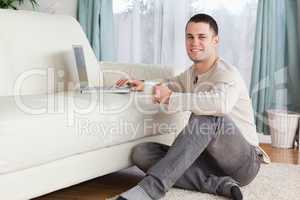 Man sitting on a carpet with a cup of tea using a laptop