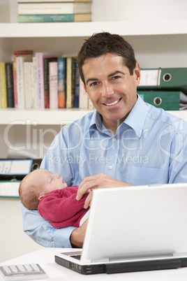 Father With Newborn Baby Working From Home Using Laptop