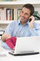 Father With Newborn Baby Working From Home Using Laptop