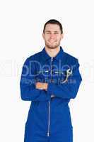 Smiling young mechanic in boiler suit with wrench and arms folde