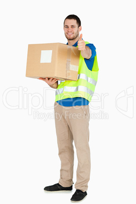 Smiling young delivery man giving thumb up while holding parcel