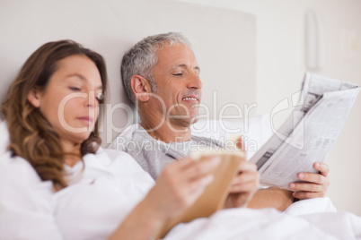 Happy woman reading a book while her husband is reading a newspa