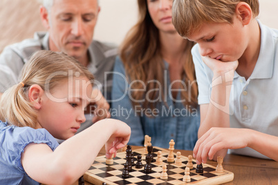 Focused siblings playing chess in front of their parents