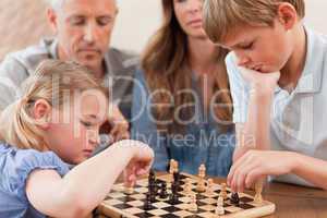Focused siblings playing chess in front of their parents
