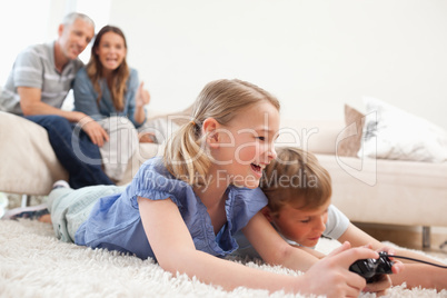 Siblings playing video games with their parents on the backgroun