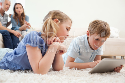 Siblings using a tablet computer with their parents on the backg