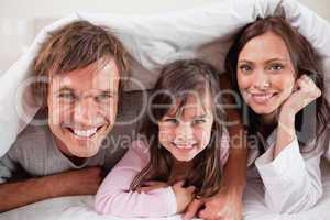 Happy parents lying under a duvet with their daughter