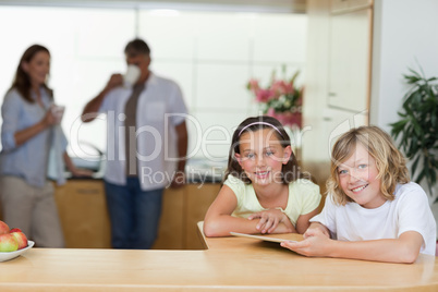 Siblings using tablet in the kitchen with parents behind them