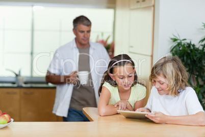 Children using tablet in the kitchen with father behind them
