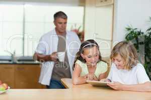 Children using tablet in the kitchen with father behind them