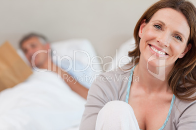 Smiling mature woman on bed with reading husband behind her