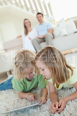 Siblings using tablet on the carpet with parents behind them