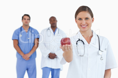 Doctor offering apple with colleagues behind her