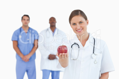 Smiling doctor offering apple with colleagues behind her