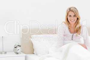 Woman sitting in bed smiling and looking forward with alarm cloc
